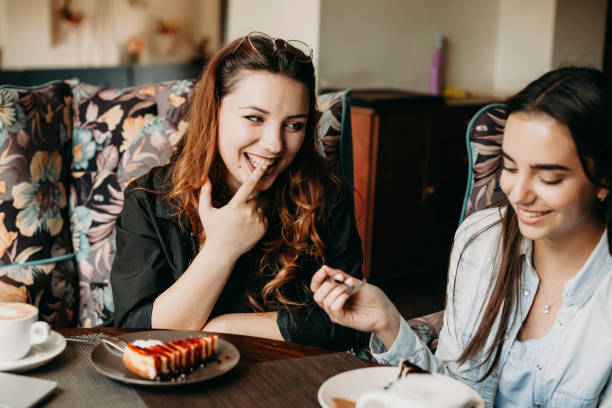 istock Beautiful red hair woman laughing while tasting a cake with her finger while looking at her girlfriend sitting in a coffee shop. 1149618336