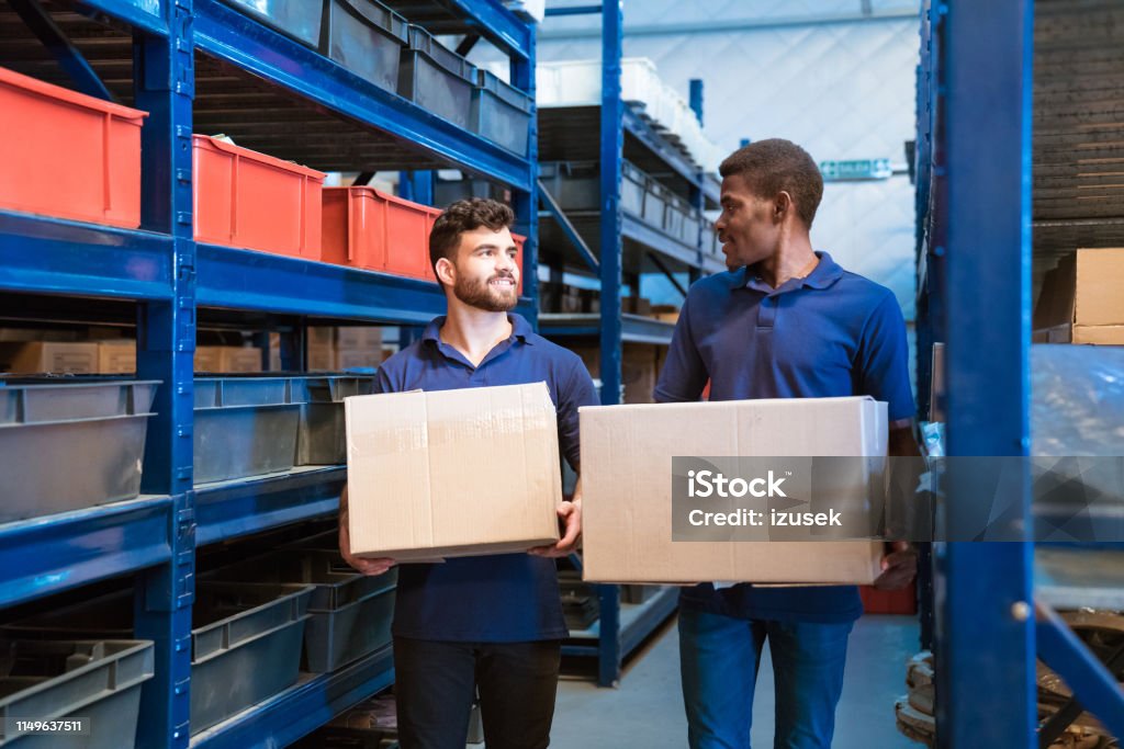 Workers carrying cardboard boxes in factory Workers carrying cardboard boxes by racks. Colleagues are standing in distribution warehouse. They are wearing uniform. Polo Shirt Stock Photo
