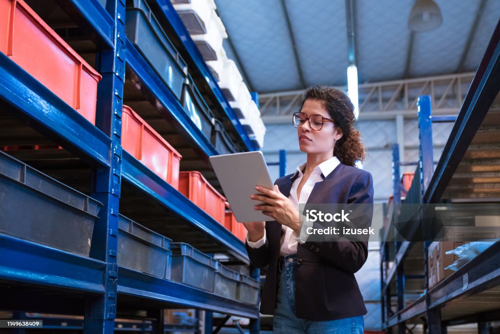 Female inspector using digital tablet by racks Female manager using digital tablet by racks. Inspector is on visit in manufacturing industry. She is standing at distribution warehouse. Manufacturing Stock Photo