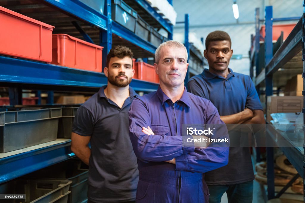 Portrait of confident foreman with coworkers Portrait of confident foreman with coworkers in factory. Manual workers are wearing uniform while standing in distribution warehouse. They are working together. Manufacturing Occupation Stock Photo