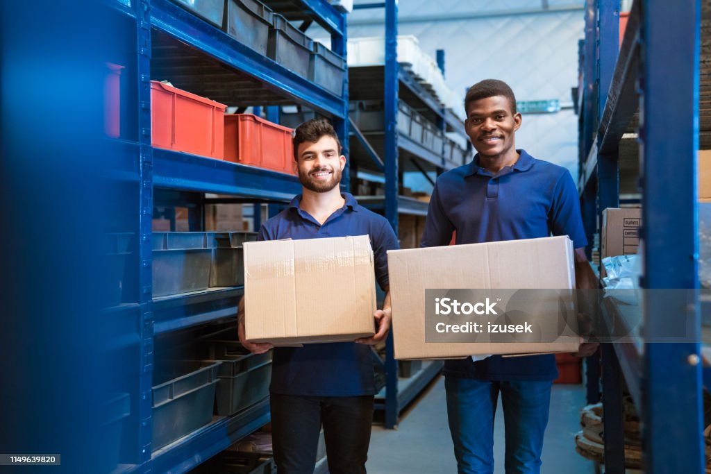 Portrait of smiling warehouse workers with boxes Portrait of smiling male warehouse workers carrying cardboard boxes. Colleagues are standing in distribution workshop. They are working together. Cardboard Box Stock Photo