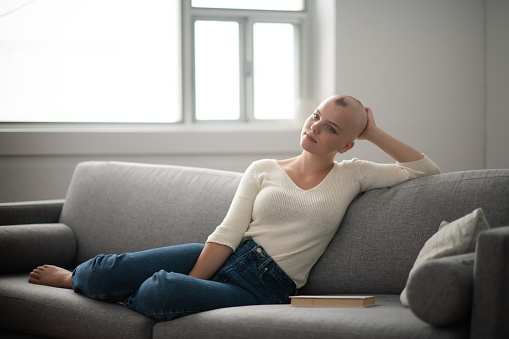 A woman is indoors in her living room. Her head is shaved due to chemotherapy. She is sitting on the couch and smiling.