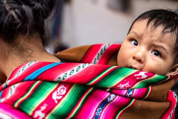 Cusco, Peru - March 31 2019: Peruvian indigenous mother carries her baby son on her back with traditional inca fabric. Native poor baby at local market.