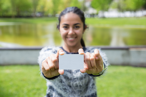 Smiling young woman showing blank plastic card in city park. Blurred pretty lady wearing sweater and standing with blurred green view in background. Introduction concept. Front view.