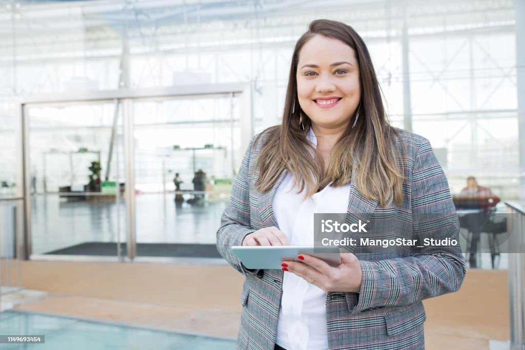 Positive lady with tablet posing in business center Positive lady with tablet posing in business center. Fat young woman in office jacket holding tablet and smiling at camera. Digital communication concept Overweight Stock Photo