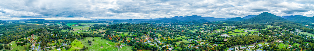 Ultra wide aerial panorama of scenic rural landscape with houses surrounded by trees, grasslands, and mountains.