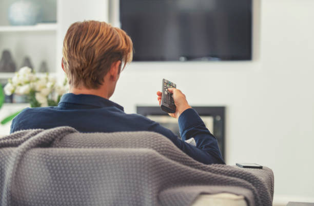 Rear view of a man watching television. Rear view of a man watching television. He is sitting on a sofa and the TV can be seen in the background. He is holding a remote control. back of head photos stock pictures, royalty-free photos & images