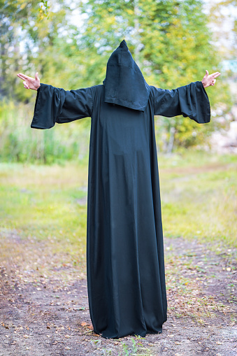 A tall man in a black suit with a hood spread his hands to the sides, showing gestures. Vertical photography