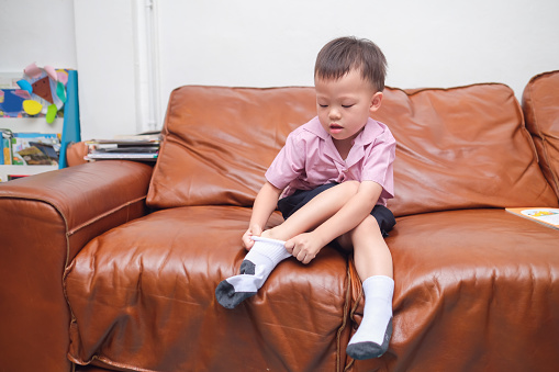 Cute little Asian 2 - 3 years old toddler boy child sitting on sofa in living room concentrate on putting on his own socks, Encourage Self-Help Skills in Children, Develop Confidence concept