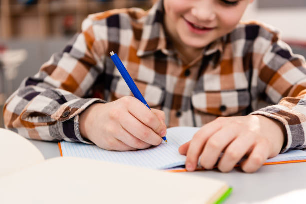 istock Partial view of smiling pupil in checkered shirt writing in notebook during lesson 1149728964