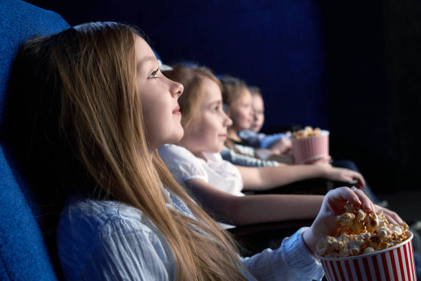 View from side of beautiful little girl sitting in cinema and watching interesting film. Young spectator resting with friends and eating popcorn. Concept of having fun and entertainment. stock photo