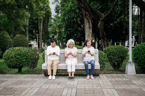 Three female, senior Taiwanese friends sitting on a bench at the park and looking at their smartphones together.