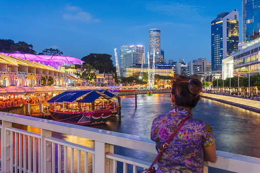 A woman stands by the railing of Read Bridge looking towards Clarke Quay, a historical quay popular for its nightlife with many restaurants and pubs.