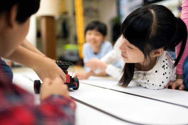 istock Young elementary girl watches as her classmate shows her how the robotic car operates 1149754993