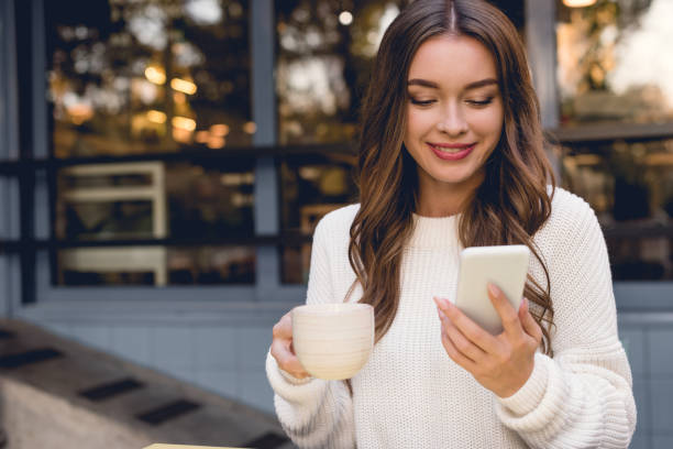 istock cheerful young woman looking at smartphone while holding cup 1149759767