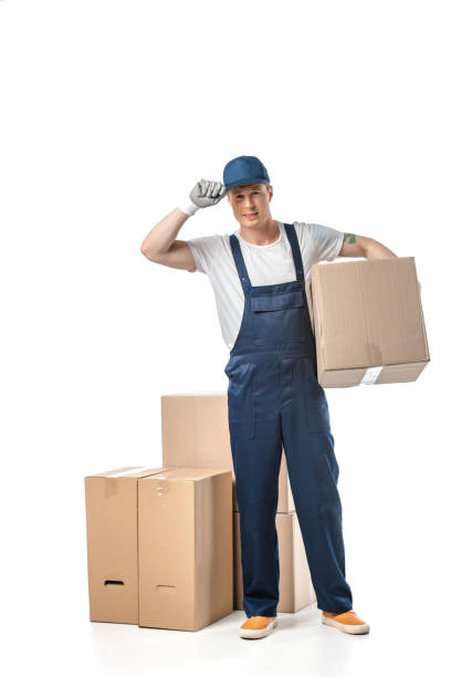 istock handsome mover in uniform adjusting hat and looking at camera while carrying cardboard box isolated on white 1149766609