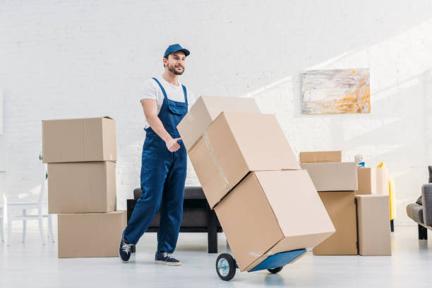 mover in uniform transporting cardboard boxes on hand truck in apartment stock photo