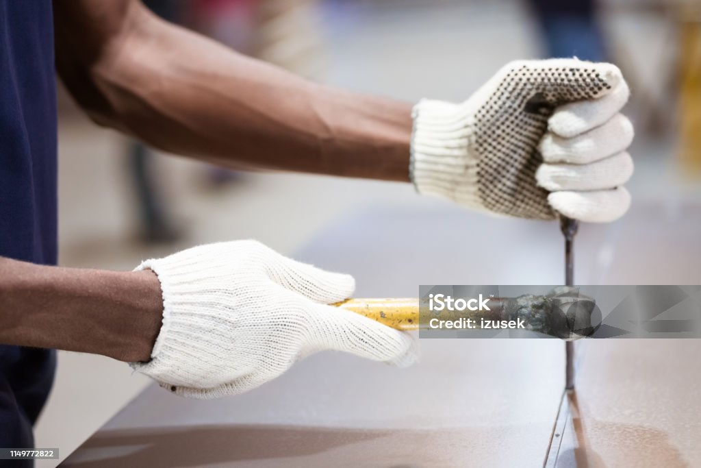 Midsection of engineer hammering machine part Midsection of male apprentice hammering on metal sheet. Engineer is wearing gloves while using work tool. He is working in factory. Manufacturing Occupation Stock Photo
