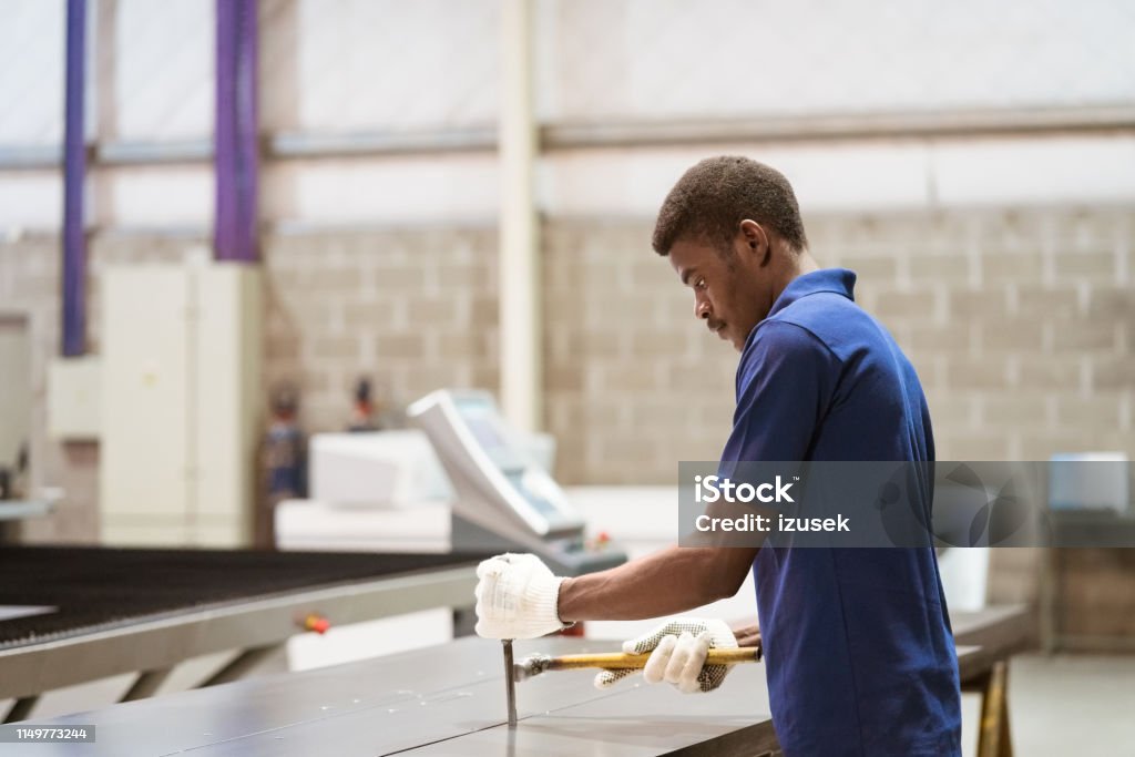 Young engineer working on metal sheet at industry Side view of young engineer working on metal sheet. Male apprentice is hammering machine part. He is standing in industry. Holding Stock Photo