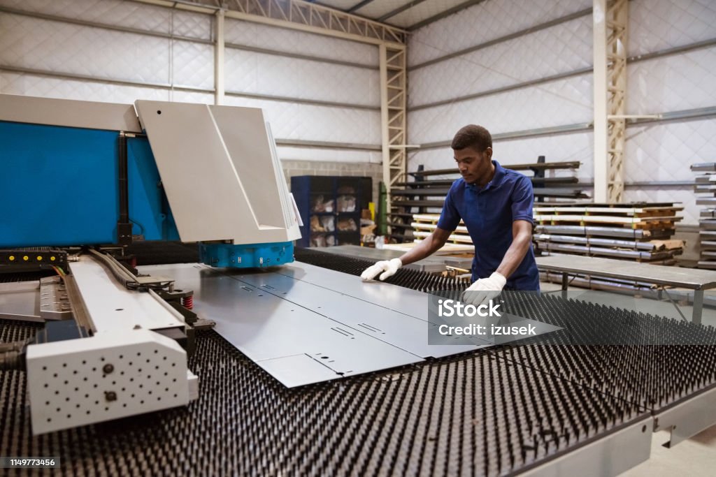 Young trainee working on puller machine in factory Male engineer adjusting metal sheet on puller machine. Young worker is wearing uniform in manufacturing industry. He is working in factory. Factory Stock Photo