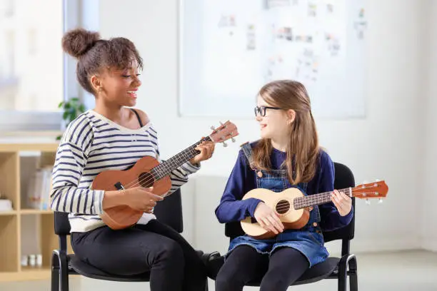Smiling multi-ethnic females playing ukulele in class. Instructor teaching music to pre-adolescent student. They are sitting in classroom at education building.