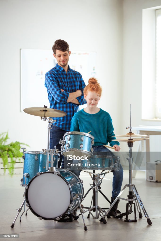 Teacher looking at drummer practicing in classroom Confident teacher looking at drummer practicing in classroom. Female student is playing drums during training class. They are at conservatory. 12-13 Years Stock Photo