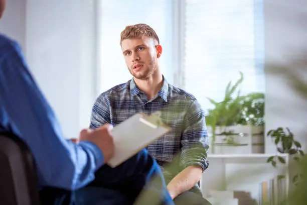 University student sharing his problems with therapist. Young depressed male is sitting with mental health professional. They are in lecture hall.