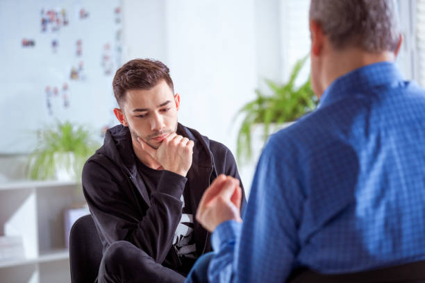 Male student listening to therapist in session Young man listening to therapist. Mental health professional discussing with student. They are in therapy session at lecture hall. information overload photos stock pictures, royalty-free photos & images