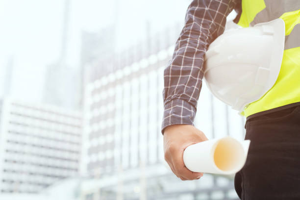 istock Close up front view of engineering male construction worker stand holding safety white helmet and wear reflective clothing for the safety of the work operation. outdoor of building background. 1149823675