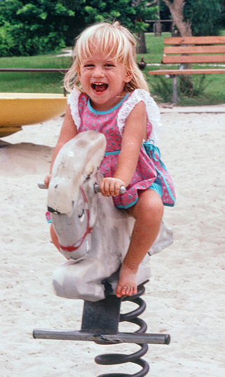 A female blonde toddler is the main interest in this vertical image. She is riding a springer horse on a playground.  She has a natural look of laughter. The daylight was bright, but no direct sun.