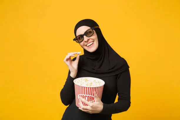 Muslim woman in hijab black clothes 3d imax glasses watch movie film hold bucket popcorn, eat pop corn isolated on yellow wall background studio portrait. People lifestyle concept. Mock up copy space