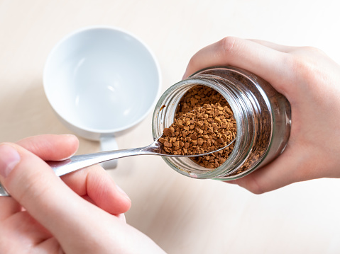 hand spooning up instant coffee from glass jar close up over cup on light brown table