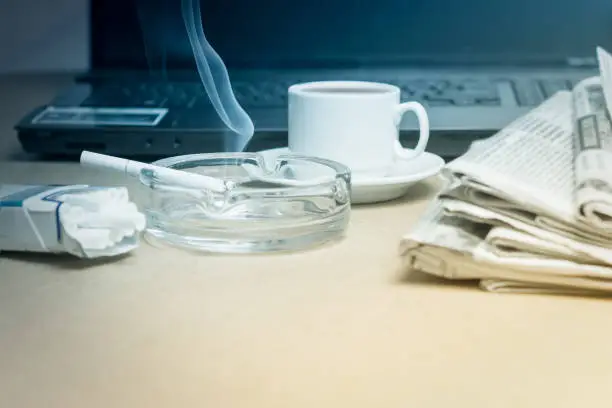 Photo of The concept of news. A folded stack of Newspapers on the laptop, a Cup of coffee, a Smoking cigarette in the ashtray. Office worker's Desk.