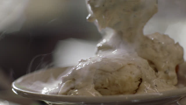 SLO MO. CU. A professional chef scoops steaming hot sausage gravy onto two biscuits at an authentic diner