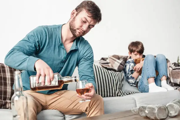 Boy and father alcoholic at home man pouring whiskey into glass concentrated while son sitting on sofa hugging bear scared