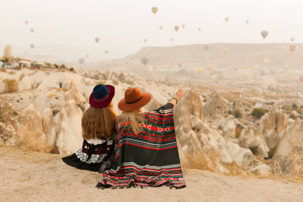 istock Traveling women wearing authentic boho chic style poncho, sweeter and hats looking on air ballons in sky in Cappadocia valley. Travel and wanderlust concept. Copy space background. 1149955315