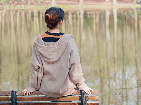 Rear view portrait of a beautiful young Chinese girl sit on park bench and face peaceful lake, enjoy sunny afternoon.