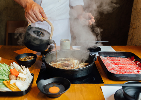 Chef is pouring soy sauce on stir fried vegetables such as onion, cabbage, scallion and tofu and more in hot pot with steam before boiling Wagyu beef.