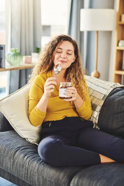 Shot of a young woman eating chocolate from a jar while relaxing on the sofa at home