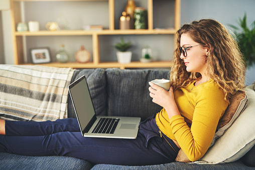 Shot of a young woman using a laptop and having coffee on the sofa at home