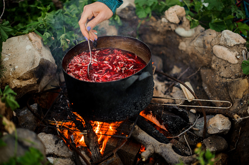 Man stirs food in a black metal pan on campfire among rocks and grass