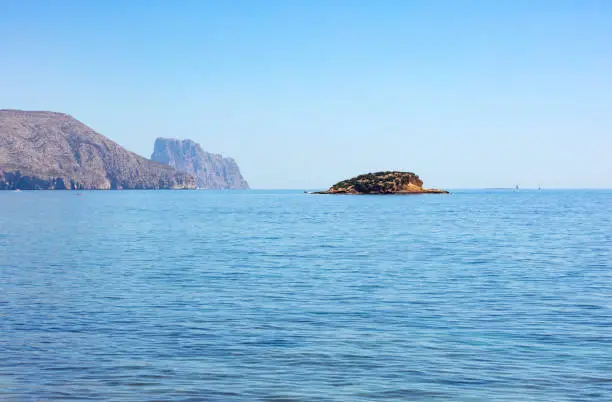 the island of La Olla in Altea, Spain, with the Ifach rock in the background in a blue mediterranean sea, calm and cristal clear