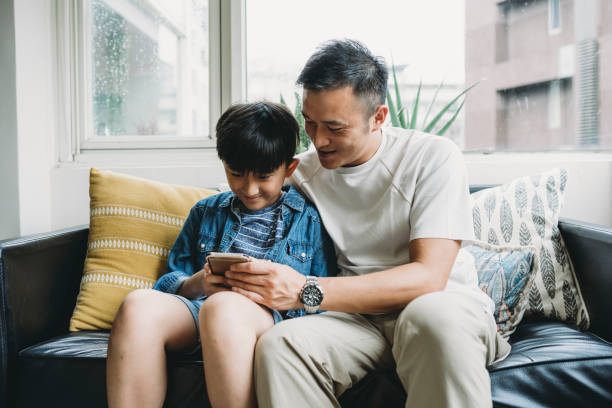 отец и сын, используя мобильный телефон вместе, сидя на диване дома - video game family child playful стоковые фото и изображения