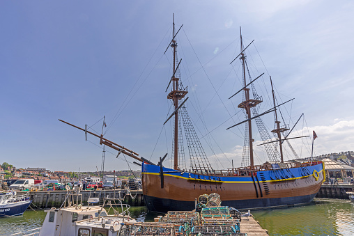 A replica of HMS Endeavour moored in Whitby Harbour. The colourful hull is in the foreground with masts projecting upwards.  A blue sky is above.