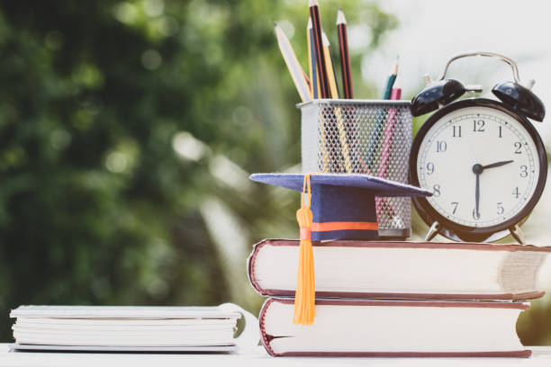 istock Graduate or Education knowledge study abroad concept : Graduation cap on textbook with pencil box and retro clock on desk, blur green light background. Back to School 1150049247