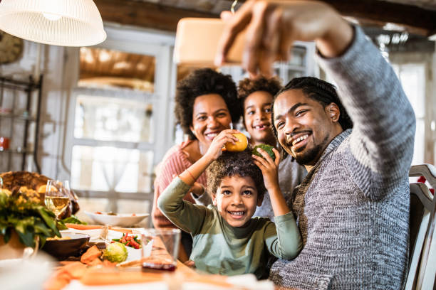 Happy black family taking a selfie with cell phone in dining room. Happy African American family having fun while taking a selfie during Thanksgiving meal at home. Focus is on father and son. dining table photos stock pictures, royalty-free photos & images