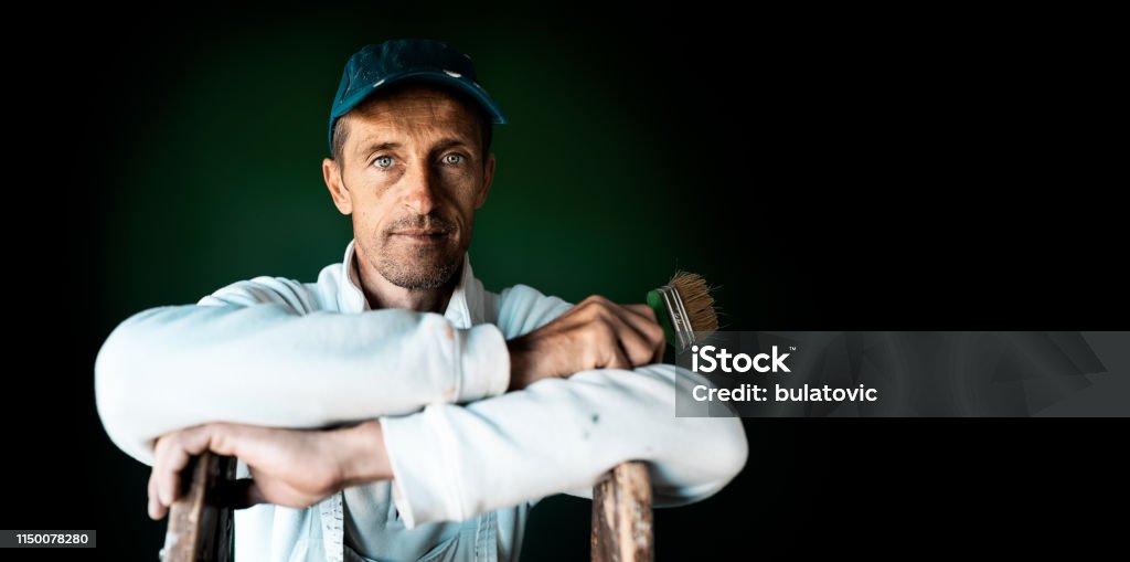 Home painter posing. Commercial. Concept. Portrait of a experienced home painter posing with a painting brush in apartment in front of dark wall House Painter Stock Photo