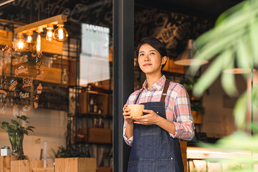A female entrepreneur standing in front of her new business, a small coffee shop in Taipei. She is holding a cup of coffee, looking off into the distance.