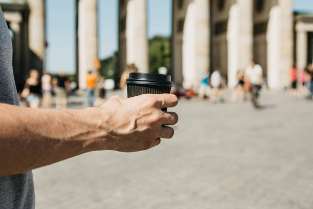 A person holds a disposable cup with coffee or another drink on the background of the Brandenburg Gate in Berlin.