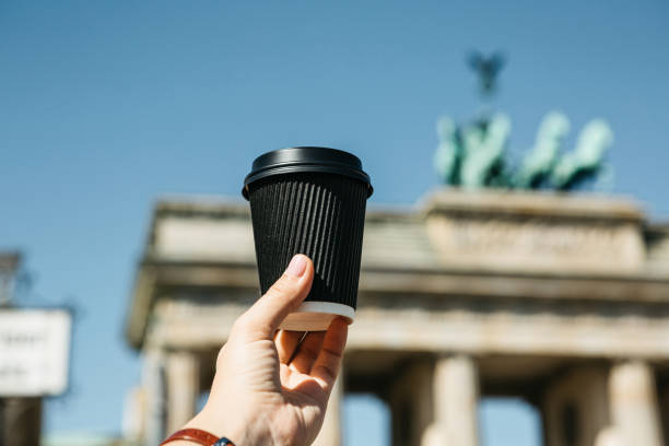 A person holds a disposable cup with coffee or another drink on the background of the Brandenburg Gate in Berlin.